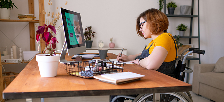 Young woman in wheelchair making notes in front of computer monitor while sitting by desk at home during remote lesson